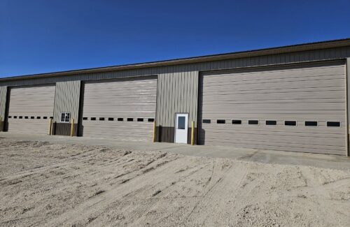 Large beige metal warehouse with three closed overhead doors on a rustic dirt lot.
