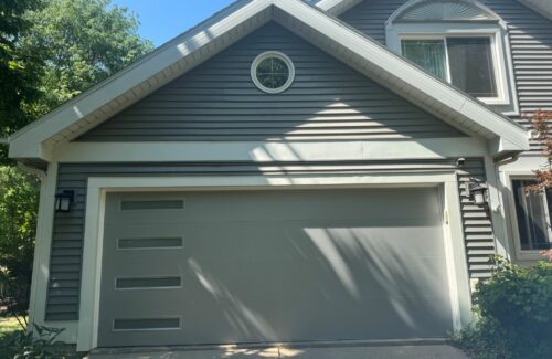 Modern gray garage with horizontal siding, a circular window, and leafy shadows.