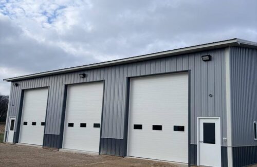 Large gray industrial garage with three white roll-up doors and an entry door on unpaved ground.