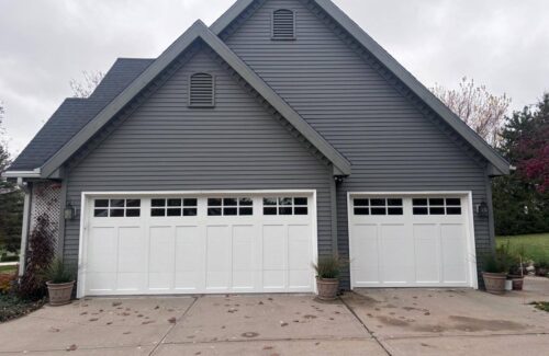 Gray house exterior with potted plants and two white garage doors featuring small square windows.