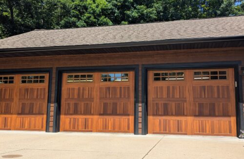 Three wooden garage doors framed by a dark brown structure and surrounded by lush green trees.
