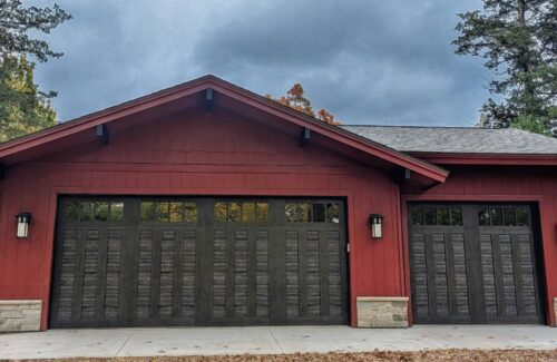Rustic red garage with stone accents and two dark wood-paneled doors surrounded by tall trees.