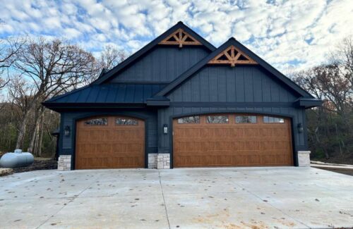 Modern dark blue house with wooden double garage doors and a concrete driveway among trees.